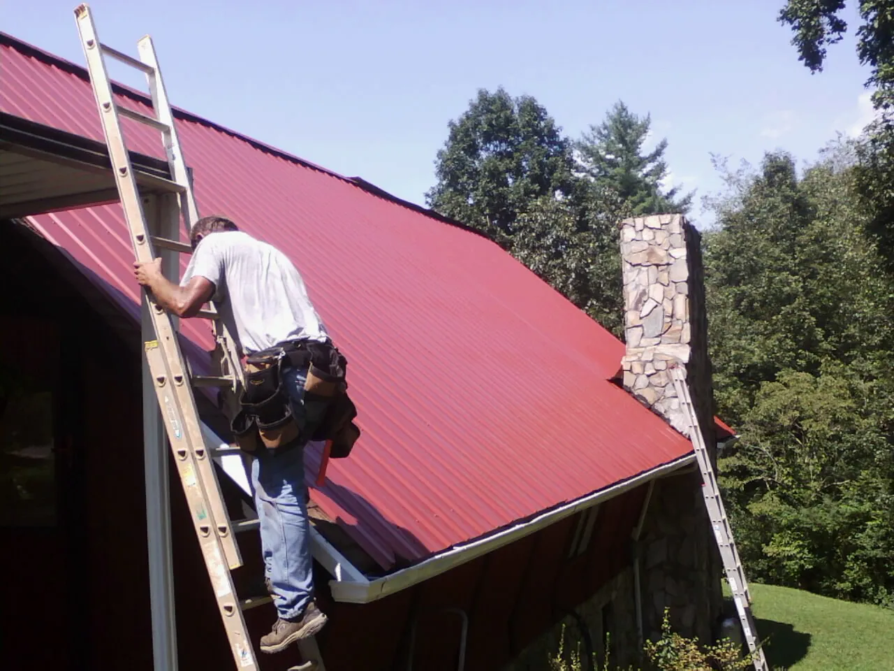 Roofer on a ladder beside a red metal roof and stone chimney — Soffit & Fascia Repair by Roofing Brothers in Corry